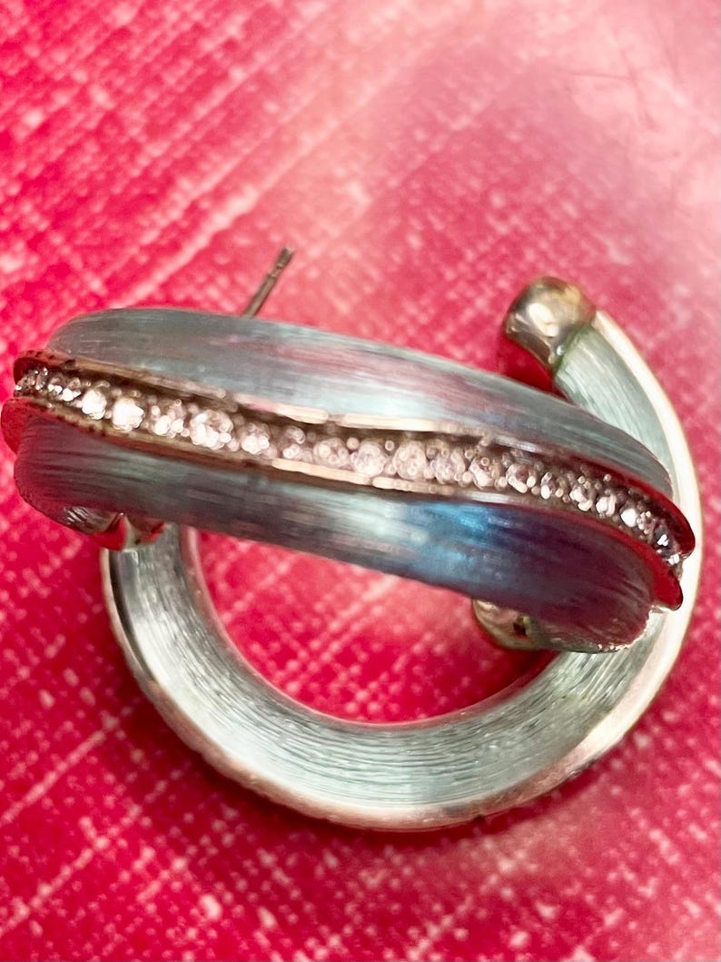 May include: A silver hoop earring with a blue and silver enamel finish. The earring has a row of small, clear crystals. The background is a bright red. The earring is a stylish accessory.