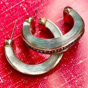 May include: Silver-coloured hoop earrings with a light blue, textured finish. The earrings feature a row of small, pink gemstones along the outer edge. Set against a red background.