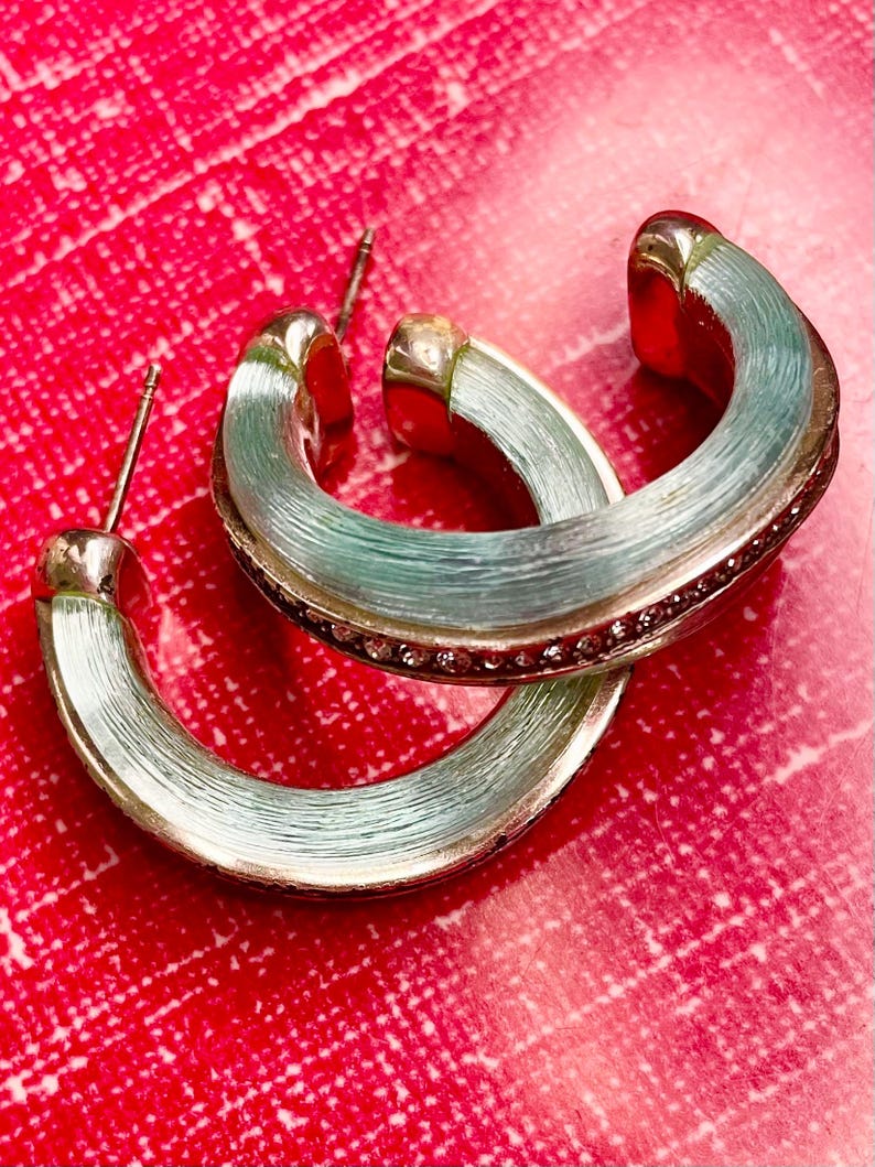 May include: Silver-coloured hoop earrings with a light blue, textured finish. The earrings feature a row of small, pink gemstones along the outer edge. Set against a red background.