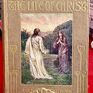 以下が含まれることがあります： 「Stories From The Life of Christ Told To Children」と題されたヴィンテージ本。表紙には、山と夕焼けのある風景の中に2人の人物が描かれたイラストが描かれています。本は、金色の文字と装飾的な要素が施されたベージュ色の表紙です。