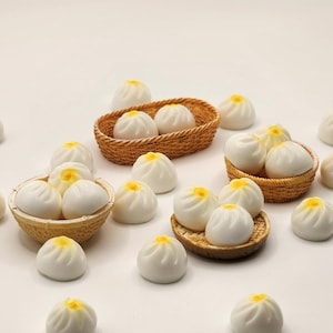 May include: Miniature white bao buns with yellow centers are arranged on a white surface, some in small woven baskets. These small, detailed food miniatures are perfect for dollhouses or craft projects.