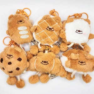 May include: A collection of plush keychains in various designs, including a cookie, waffle, and bread shapes. Each keychain is brown with a smiling face and attached to a gold-colored clasp.
