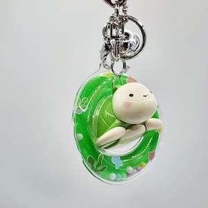 May include: A keychain featuring a small, white-faced turtle figurine inside a clear, green-ringed float. The float has a green border with the text "Marine Life" and is filled with colorful beads and small star-shaped confetti. The keychain has a silver clasp.