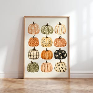 May include: A framed art print featuring a grid of twelve pumpkins in various colours and patterns, including floral, plaid, leopard print, and solid orange, green, and black designs, all set against a light cream-coloured background.