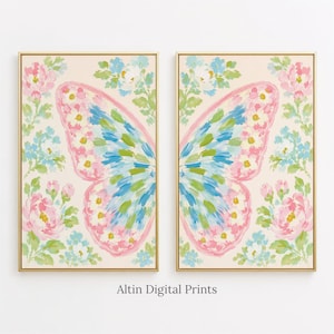 Op de afbeelding: Twee rechthoekige wandkunstprints in gouden lijsten met een gespleten vlindervleugelontwerp. De vleugels zijn geschilderd met roze, blauwe en groene penseelstreken, omringd door trossen roze en lichtblauwe bloemen op een crèmekleurige achtergrond.