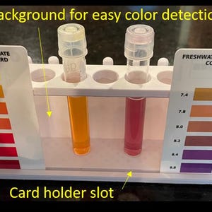 May include: A white plastic test tube holder with two test tubes containing orange and pink liquids. The holder has slots for color cards labeled "FRESHWATER NITRATE" and "FRESHWATER HIGH RANGE pH". The text "White background for easy color detection" is visible.