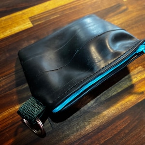 Puede incluir: Bolsa rectangular negra hecha de material de neumático reciclado. La bolsa presenta una cremallera azul brillante y un lazo de tela verde con un anillo de metal. La bolsa está sobre una superficie de madera.