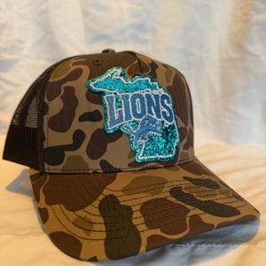 May include: A camouflage baseball cap with a mesh back. The front of the cap features a teal and blue glitter patch in the shape of the state of Michigan with the word "LIONS" in blue lettering.