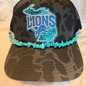 May include: A camouflage-patterned trucker hat with a black mesh back. The front features a teal glitter patch of the state of Michigan with the word "LIONS" and a turquoise stone bead trim.