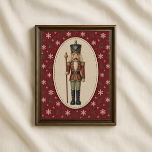 Op de afbeelding: Een ingelijst wandkunstwerk met een centrale ovale illustratie van een notenkraker-soldaat in een rood en goud uniform die een staf vasthoudt. De achtergrond is een dieprood patroon met witte sneeuwvlokken en kleine groene hulstblaadjes.