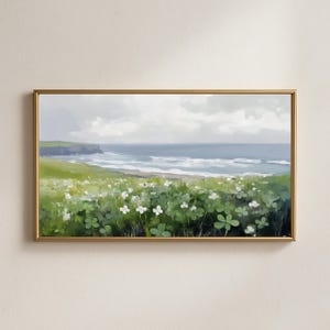 May include: A framed landscape painting depicts a coastal scene. The artwork features a green field of clovers and white flowers in the foreground, transitioning to a blue ocean under a cloudy sky. The painting is encased in a gold frame.