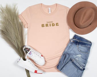 Puede incluir: Una composición plana con una camiseta color melocotón con la inscripción dorada "team BRIDE", un sombrero marrón, vaqueros de mezclilla descoloridos, zapatillas blancas con estampado de leopardo y hierba seca decorativa. Los artículos están dispuestos sobre una superficie blanca.