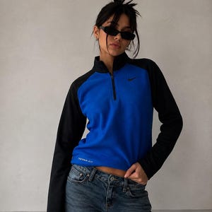 Könnte beinhalten: Ein blau-schwarzer Langarm-Pullover mit Stehkragen und kurzem Reißverschluss. Das Kleidungsstück hat ein schwarzes Nike-Logo auf der linken Brust und den Schriftzug THERMA-FIT in Weiß nahe dem unteren rechten Saum.