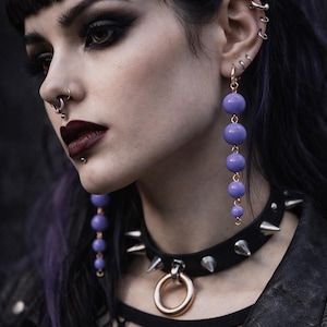 Può includere: Una persona con capelli scuri e trucco indossa un choker nero con borchie argentate e un anello dorato. Indossa anche orecchini pendenti lunghi con perline viola. L'estetica generale è gotica e audace.