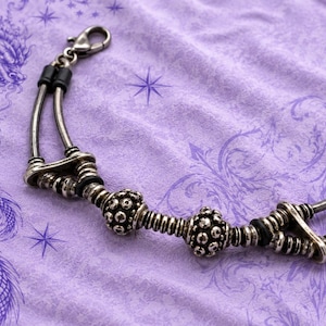 Può includere: Un bracciale color argento caratterizzato da due barre metalliche curve parallele collegate da perline decorative testurizzate e piccoli anelli in gomma nera. Il gioiello include una chiusura a moschettone e perline metalliche arrotondate.