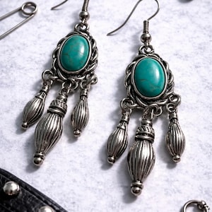 Può includere: Un paio di orecchini pendenti color argento con pietre centrali ovali turchesi e tre pendenti decorativi a forma di goccia. Presentati su una superficie chiara, insieme ad altri accessori argentati.