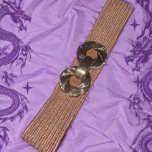 Può includere: Una cintura larga intrecciata color beige con una grande fibbia circolare in metallo dorato dal design annodato e testurizzato. La cintura è realizzata in materiale intrecciato ed è posizionata in diagonale, mettendo in risalto la chiusura.