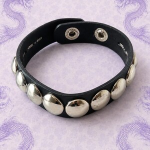 Può includere: Un braccialetto nero caratterizzato da una fila di borchie circolari color argento, lisce e lucide, sulla superficie esterna. Il cinturino include due bottoni a pressione in metallo color argento sul lato interno per chiudere il bracciale.