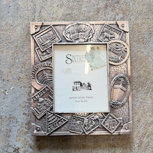 May include: A rectangular pewter photo frame featuring a textured background and embossed travel-themed motifs including landmarks from Europe, Canada, India, and the Orient. The center insert displays the text Sixtren, pewter photo frame, four by six.