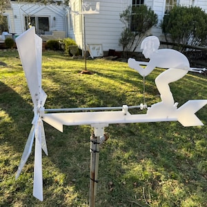 May include: A white metal weather vane depicting a skier, mounted on a pole. The design includes a directional arrow and a small windmill. The weather vane is set in a grassy garden with a house in the background.