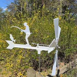 May include: A white metal weathervane featuring a fisherman silhouette with a fishing rod and a fish. The weathervane has a propeller and is mounted on a metal pole. The background includes a wooden fence and yellow flowering bushes.