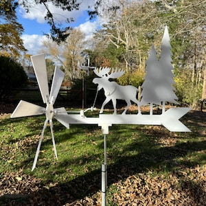 May include: A white metal weather vane featuring a moose, pine trees, and a directional arrow. The vane also includes a small windmill. The design is set in a grassy yard with trees and a blue sky in the background.