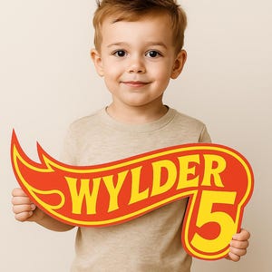May include: A child holds a red and yellow sign with the text "WYLDER 5". The sign has a flame design, possibly for a birthday. The child wears a beige top and blue jeans. The image is well-lit and the child is smiling.
