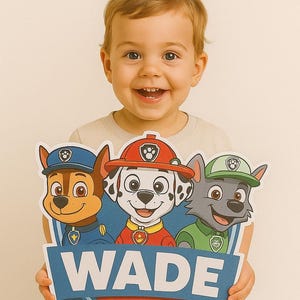 Puede incluir: Un niño sonriente sostiene un letrero colorido con personajes de Paw Patrol. El letrero muestra el nombre "WADE" en letras azules, con un diseño en forma de hueso. Los personajes de dibujos animados visten sus uniformes.