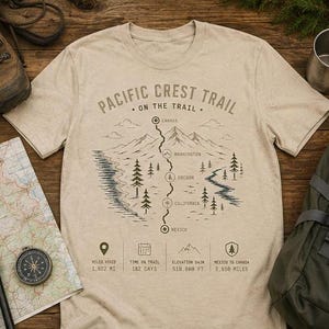 May include: A beige short-sleeve t-shirt featuring a dark blue graphic of the Pacific Crest Trail. The design includes a mountain range, pine trees, a winding trail path, and text labels for Canada, Washington, Oregon, California, and Mexico.