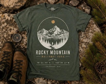 May include: Green t-shirt with a circular graphic of a mountain range, lake, and trees, with the text "ROCKY MOUNTAIN NATIONAL PARK". Coordinates and "COLORADO, USA" are also printed on the shirt. Brown hiking boots and a compass are next to the shirt.