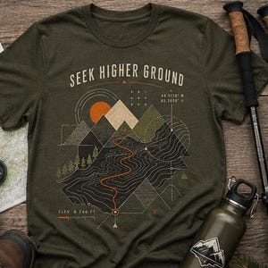May include: An olive green short-sleeve t-shirt featuring a graphic print of mountains, a winding trail, and the text "SEEK HIGHER GROUND" with coordinates and elevation data. The design includes geometric lines, a sun, and small pine trees.