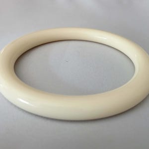 Puede incluir: Un brazalete circular de color crema. La superficie lisa y brillante refleja la luz, resaltando su forma redondeada. Este accesorio simple y elegante es adecuado para el uso diario.