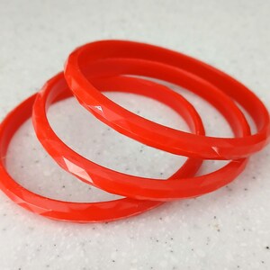 Könnte beinhalten: Drei leuchtend rote Armreifen mit facettiertem Design liegen auf einer gesprenkelten weißen Oberfläche. Die Armreifen bestehen aus einem festen Material und haben ein glänzendes Finish. Der Schmuck ist ein schlichtes, aber auffälliges Accessoire.