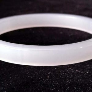 May include: A smooth, solid white bangle bracelet with a rounded exterior profile and a polished, semi-translucent finish. The circular jewelry piece features a consistent thickness throughout its entire circumference, presenting a clean, simple design.