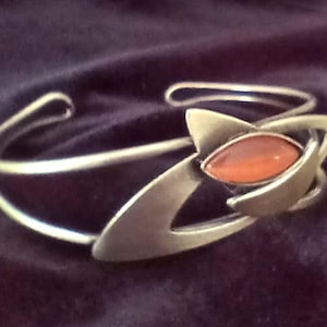 May include: A silver-toned metal cuff bracelet featuring a double-band design and a central focal point with an elongated, marquise-shaped orange stone set within a smooth, abstract geometric frame that curves around the wrist.