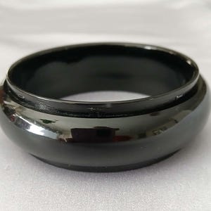 Puede incluir: Un cuenco negro brillante, de perfil bajo, con una base ancha y redondeada y un borde ligeramente elevado. La superficie del cuenco refleja la luz, creando reflejos y sombras. El diseño es simple y moderno, adecuado para diversos usos decorativos o funcionales.
