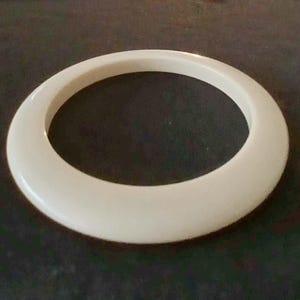 Puede incluir: Un brazalete circular de color crema. El brazalete está hecho de un material sólido y liso y tiene una forma redondeada. El brazalete es un accesorio simple y elegante.