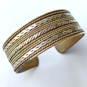 Könnte beinhalten: Ein breites Armband mit Gold-, Silber- und Kupfertönen. Das Armband hat komplizierte geflochtene und perlenbesetzte Muster, die ein strukturiertes, geschichtetes Aussehen erzeugen. Das offene Design ermöglicht ein einfaches Tragen. Ca. 4 cm breit.