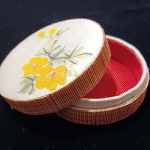 May include: A round jewelry box with a bamboo-style exterior and a white lid. The lid features a floral design with yellow flowers and green leaves. The interior is lined with red fabric.