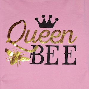 May include: Pink fabric with the words "Queen BEE" in gold glitter and black lettering. A black crown sits above the word "Queen", and a gold glitter bee is below the word "Queen".