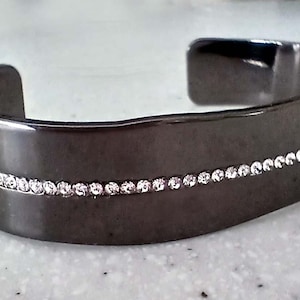 May include: A dark gray cuff bracelet with a row of small, clear crystals. The bracelet has a smooth, polished finish and a simple, elegant design. This jewelry item is suitable for everyday wear or special occasions.