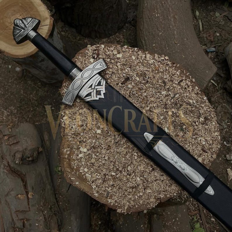 Puede incluir: Una espada de estilo vikingo con empu&ntilde;adura y vaina negras. La espada presenta una guarda y pomo plateados con intrincados dise&ntilde;os de nudos celtas. La vaina tiene una contera plateada. La espada descansa sobre una superficie de madera.