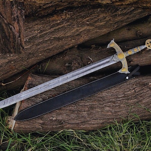 May include: A steel sword with a patterned blade and an ornate gold and silver-toned hilt featuring a cross design on the pommel. The sword is accompanied by a black leather scabbard with a silver metal tip and a strap for securing the handle.