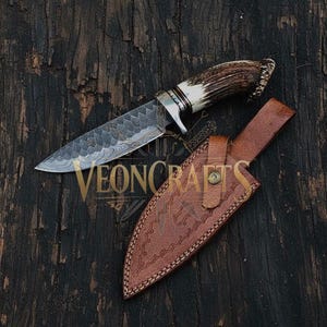 Puede incluir: Un cuchillo de caza artesanal con una hoja de acero estampada y un mango de asta y hueso. El cuchillo descansa sobre una superficie de madera oscura junto a una funda de cuero marrón. El texto "VEONCRAFTS" es visible.