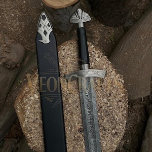 Puede incluir: Una espada vikinga con una hoja estampada y una empu&ntilde;adura plateada ornamentada. La espada tiene un mango negro y una vaina negra. La hoja presenta texto grabado. La espada descansa sobre una superficie de madera.