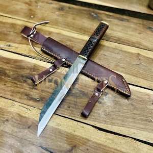 Könnte beinhalten: Ein handgefertigtes Messer mit polierter Stahlklinge und einem dekorativen Griff mit keltischem Knotenmuster. Das Messer liegt auf einer braunen Lederscheide mit passender Gürtelschlaufe. Messer und Scheide liegen auf einer Holzoberfläche.