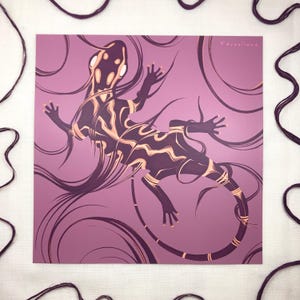 May include: A square print featuring a stylized lizard illustration in dark purple and gold against a mauve background. The lizard is surrounded by swirling lines, with the word "Feroxious" in the upper right corner.