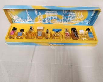 Op de afbeelding: Een verzameling miniatuur parfumflesjes in een decoratieve doos. De doos is geel en blauw, met illustraties van Parijse bezienswaardigheden. De flesjes hebben verschillende vormen en maten en zijn gevuld met amberkleurige vloeistof.