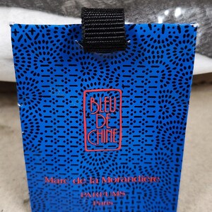 Pu&ograve; includere: Una scatola di profumo blu con un cinturino nero. La scatola presenta un motivo ripetuto di piccoli punti neri e un logo rosso che recita "BLEU DE CHINE". &Egrave; visibile anche il testo "Marc de la Morandi&egrave;re, PARFUMS Paris".