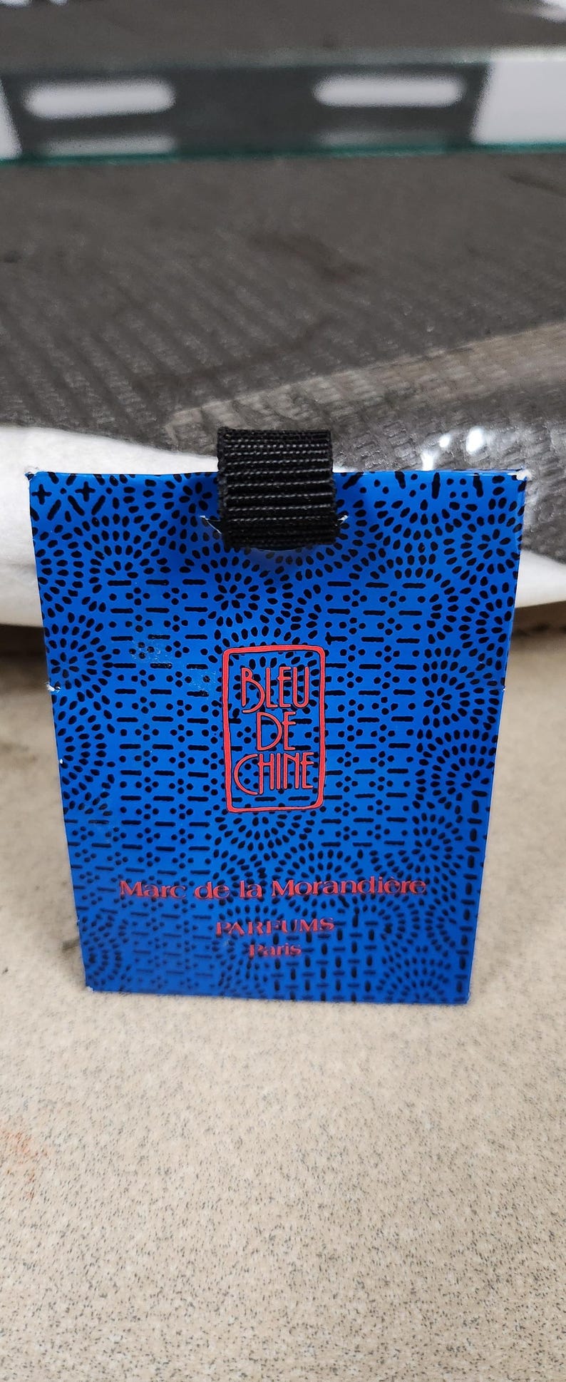 Pu&ograve; includere: Una scatola di profumo blu con un cinturino nero. La scatola presenta un motivo ripetuto di piccoli punti neri e un logo rosso che recita "BLEU DE CHINE". &Egrave; visibile anche il testo "Marc de la Morandi&egrave;re, PARFUMS Paris".